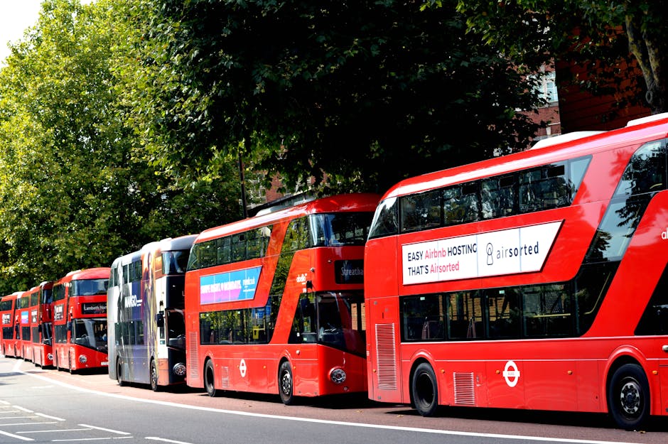 A row of red double-decker buses parked along a street in Lambeth, with the first bus in the foreground displaying an advertising banner on its side that reads 'EASY Airbnb HOSTING THAT'S Airsorted.' Behind the buses, there are green leafy trees and part of a red-brick building visible. The scene is set during daylight with clear weather, and the buses are aligned on the right side of the road, which appears to be a designated bus lane or stopping area. The image captures the urban environment associated with a typical home relocation or furniture transport process, reflecting the importance of logistical planning in house removals and moving services, as seen through the contextual inclusion of transport vehicles in the vicinity of residential streets preferred by services like Man with Van Lambeth.