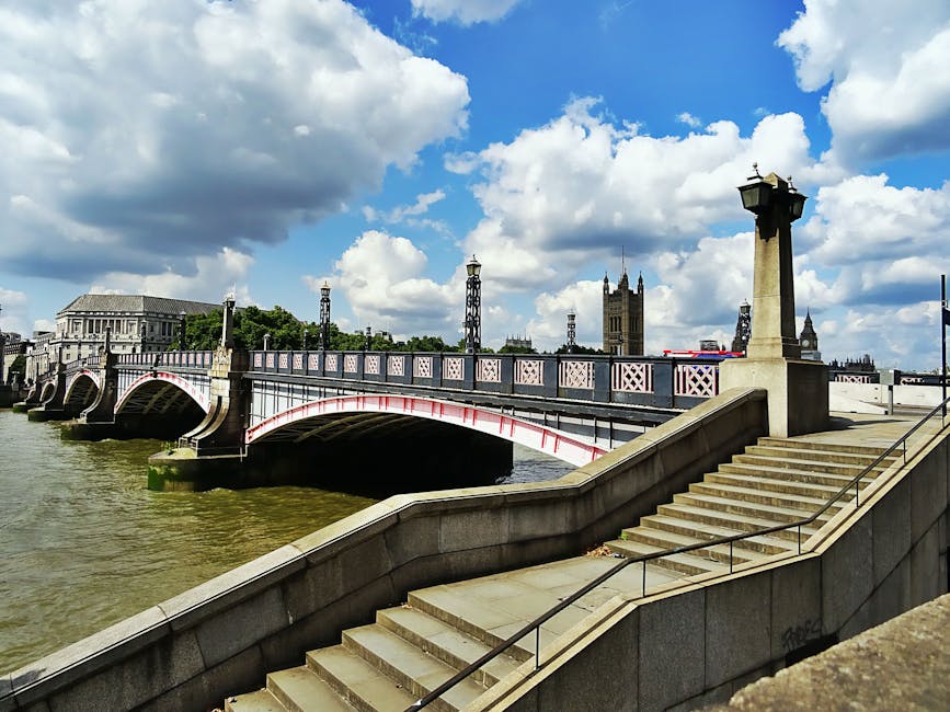 A stone staircase with metal handrails leading down from a raised promenade alongside the River Thames in London. Adjacent to the stairs, a section of pavement is visible, with the river flowing underneath a historic bridge that features spaced, ornate lampposts and decorative railings painted in pink and white. In the background, iconic tall buildings, including a clock tower resembling Westminster Palace, are visible against a partly cloudy sky with large, fluffy clouds casting soft shadows. The scene captures an outdoor view of a cityscape associated with London, highlighting the architectural and infrastructural elements typical of a riverside area. The environment appears well-lit with natural daylight, emphasizing the details of the bridge, stairs, and surrounding structures, consistent with a typical city day. This setting relates to house and furniture removals, possibly showing access points or logistics pathways for moving services, as would be overseen by companies like Man with Van Lambeth.
