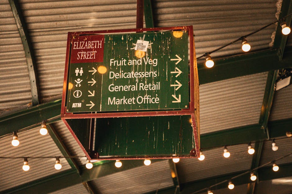 A large, rectangular green and red directional sign hanging from a ceiling with exposed green metal beams and corrugated metal roofing panels inside a covered market area. The sign displays white text and arrows guiding visitors to different sections, including 'Fruit and Veg,' 'Delicatessens,' 'General Retail,' and 'Market Office.' The sign appears weathered with visible scratches and peeling paint. Around the sign, warm white string lights are suspended, providing ambient lighting. The environment suggests an active marketplace, which may require careful planning for furniture transport or home relocation logistics handled by [COMPANY_NAME], especially in narrow street access situations like Brixton Market as discussed on the relevant page.
