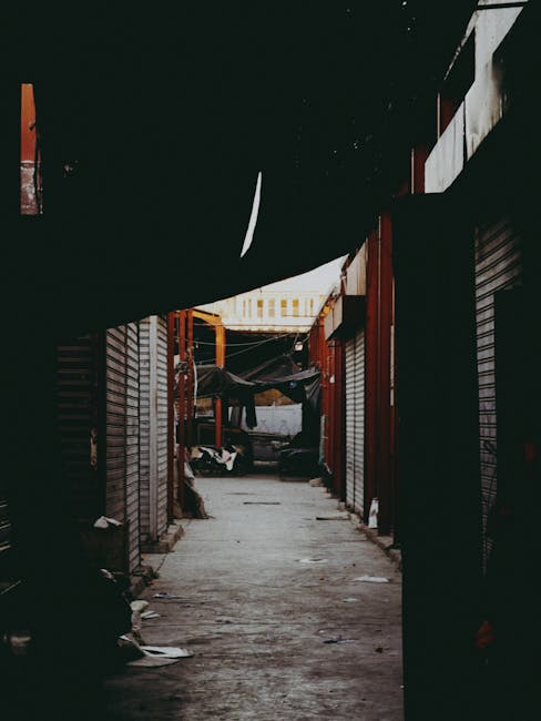 A narrow alleyway in Brixton Market with cluttered surroundings, including closed shopfronts with metal shutters on both sides and black tarps or fabrics overhead, creating a shaded environment. The ground is uneven and littered with small debris, indicating a busy or under-maintained space. At the end of the alley, a yellow crane or lifting equipment is visible, suggesting ongoing construction or a loading operation. In the foreground, part of a moving van or truck is visible, parked at the entrance to the alley, with a glimpse of furniture or packed boxes inside. This scene depicts the logistical challenges of home relocation and furniture transport through tight, urban environments, highlighting the importance of professional removals services such as those provided by Man with Van Lambeth, especially for navigating narrow street access during property moves.