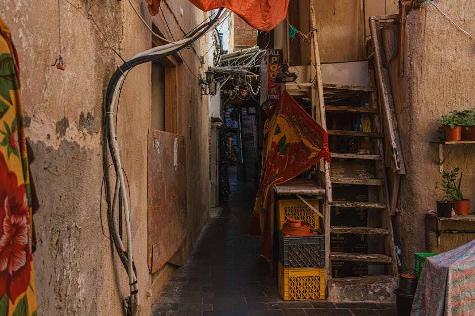 A large, rectangular green and red directional sign hanging from a ceiling with exposed green metal beams and corrugated metal roofing panels inside a covered market area. The sign displays white text and arrows guiding visitors to different sections, including 'Fruit and Veg,' 'Delicatessens,' 'General Retail,' and 'Market Office.' The sign appears weathered with visible scratches and peeling paint. Around the sign, warm white string lights are suspended, providing ambient lighting. The environment suggests an active marketplace, which may require careful planning for furniture transport or home relocation logistics handled by [COMPANY_NAME], especially in narrow street access situations like Brixton Market as discussed on the relevant page.