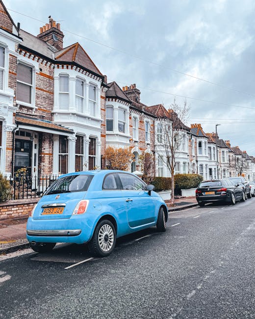 A light blue compact car parked on a residential street in Lambeth, with its rear end facing the viewer. The vehicle is situated adjacent to a row of Victorian-style terraced houses with white and brick facades, decorative bay windows, and small front gardens enclosed by low brick walls. Nearby, a black car is also parked parallel to the curb. The street is lined with leafless trees, and the pavement is clear of obstructions, indicating a typical urban setting suitable for house removals or furniture transport activities. This scene depicts an area where professional moving services, such as those provided by Man with Van Lambeth, might operate for home relocations, packing, and loading processes within residential neighbourhoods, especially considering the context of parking permits and manoeuvring in tight urban spaces.