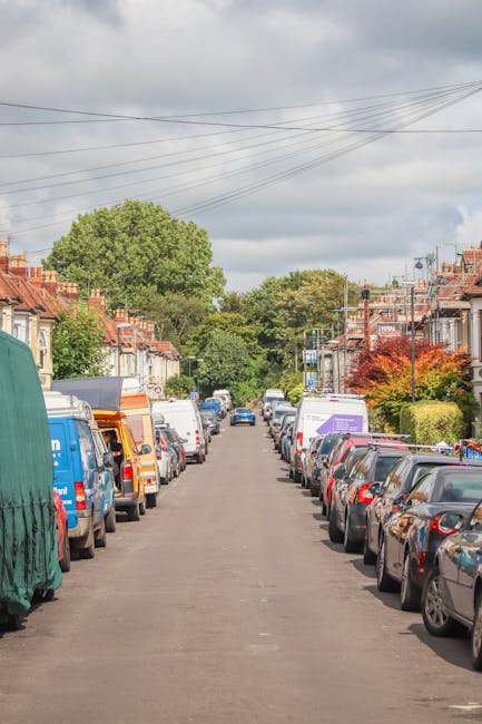A residential street in Clapham Common featuring rows of parked vehicles on both sides, including small vans, cars, and trucks, some of which are covered with protective tarps or covered with plastic sheeting. The street is lined with terraced houses with brick facades, front gardens, and rooftops, typical of London housing. In the background, there are lush green trees and a cloudy sky overhead. The scene suggests a setting where home relocation or furniture transport might be taking place, possibly involving organized loading or unloading of moving boxes and household items. Man with Van Lambeth, a house removals service, is not visible but implied to be involved in coordinating such logistics within this environment, supporting effective packing and moving procedures.