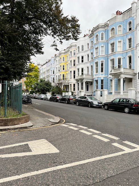 A light blue compact car parked on a residential street in Lambeth, with its rear end facing the viewer. The vehicle is situated adjacent to a row of Victorian-style terraced houses with white and brick facades, decorative bay windows, and small front gardens enclosed by low brick walls. Nearby, a black car is also parked parallel to the curb. The street is lined with leafless trees, and the pavement is clear of obstructions, indicating a typical urban setting suitable for house removals or furniture transport activities. This scene depicts an area where professional moving services, such as those provided by Man with Van Lambeth, might operate for home relocations, packing, and loading processes within residential neighbourhoods, especially considering the context of parking permits and manoeuvring in tight urban spaces.