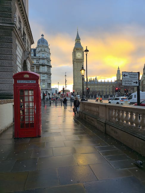A view of a London street sidewalk during twilight with the iconic Big Ben clock tower and the Houses of Parliament in the background. In the foreground, there is a traditional red British telephone box positioned near the edge of the pavement, which is paved with large rectangular stone slabs. Street lamps are illuminated, casting a warm glow on the wet surface, indicative of recent rain. Several pedestrians are seen walking along the sidewalk and crossing the road, some carrying bags. To the right, cars and a bus are visible in traffic, along with a directional sign indicating routes to Trafalgar Square and Lambeth Bridge. The scene portrays a typical London urban setting, relevant to house removals and furniture transport, as [COMPANY_NAME] might coordinate logistics for home relocation in this area.