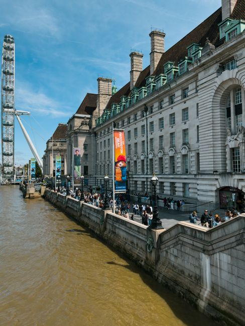 A wide view of a historic white stone building with multiple chimney stacks and teal window awnings, situated along the riverbank in Lambeth. In front of the building, a paved walkway is filled with pedestrians, some of whom are standing near the edge or walking along the promenade. Several black lampposts line the corridor, and colorful banners, including one featuring a character from a popular video game, hang from poles attached to the building. To the left, a large construction crane extends into the blue sky, indicating nearby development. The river in the foreground has a brownish hue, with gentle water movement, while a modern white suspension bridge with cables extends from the left side of the image. This setting provides a glimpse of urban waterfront activity often associated with city centre locations like Lambeth, where house removals and moves occur regularly, and the area’s architecture and transport infrastructure support efficient furniture transport and home relocation services by companies such as Man with Van Lambeth.