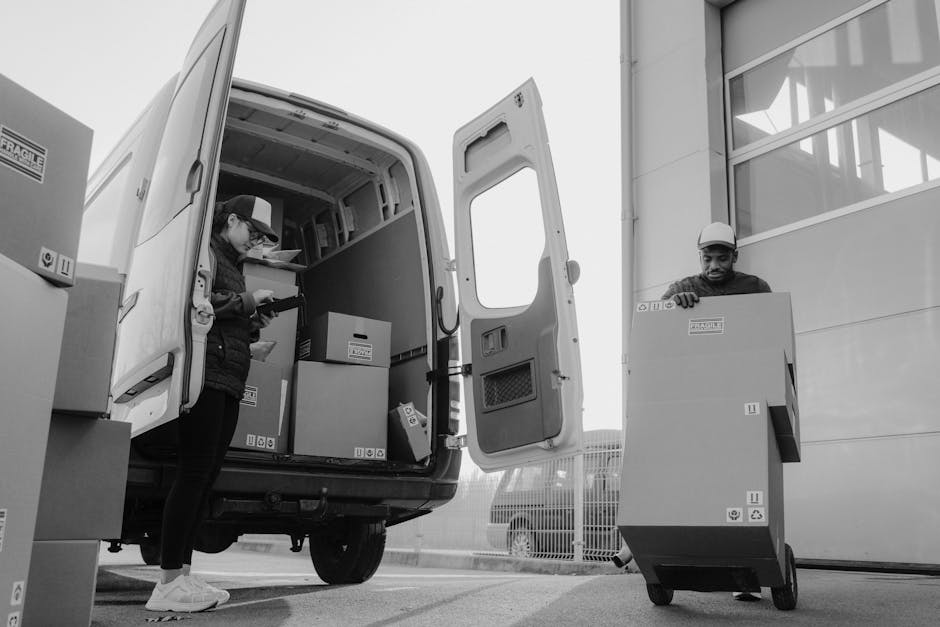A black and white photograph depicts two movers from Man with Van Lambeth engaged in a home relocation process. The scene shows them outside near a modern building with large windows and a closed roller shutter. Inside the back of a large van, which has open double doors, several cardboard moving boxes of various sizes are visible, some stacked on each other and secured with plastic wrap. The woman, wearing a cap, jacket, and sneakers, is inside the van, inspecting or preparing a box, while the man, wearing a helmet and work attire, is outside the van pushing a large cardboard box on a wheeled trolley towards the vehicle. The ground is paved, and some additional boxes are seen on the pavement near the van, highlighting the packing and loading process typical of furniture transport and house removals. This image, associated with Lambeth council permit rules for moving vans, captures the logistical aspects of loading boxes and furnishings during a professional move.