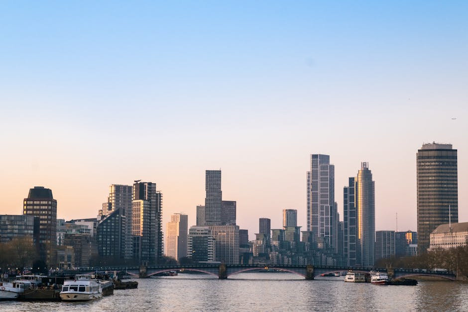 A wide-angle view of a city skyline with modern high-rise buildings situated along a river. In the foreground, there is a paved pavement with a mobile staircase and a few cardboard boxes stacked nearb