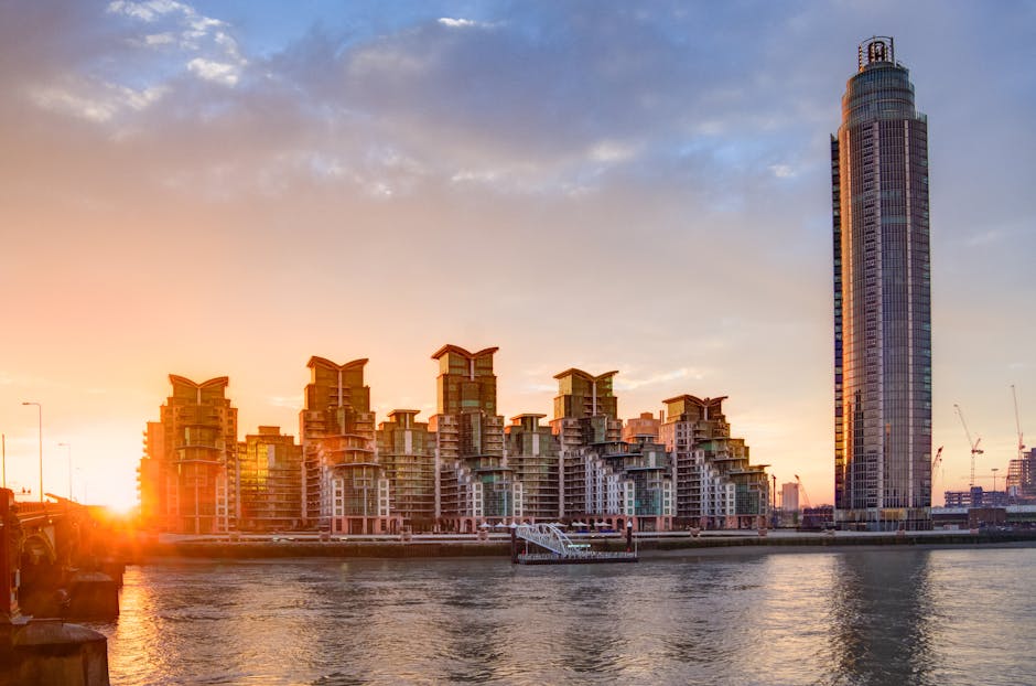 A wide-angle view of a city skyline with modern high-rise buildings situated along a river. In the foreground, there is a paved pavement with a mobile staircase and a few cardboard boxes stacked nearb