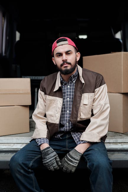 A young man with a beard and wearing a red baseball cap, a checked shirt, and a two-tone beige and brown jacket, is sitting on the edge of an open van's loading area during a home relocation. Behind him, there are several large cardboard boxes arranged on the vehicle's floor, some stacked on top of each other, ready for transport. The man is wearing grey knitted gloves with black accents, indicating preparation for handling fragile or heavy items. The vehicle is parked outdoors, and the interior is dimly lit, with black seats visible in the background. This scene likely depicts the loading process during a furniture transport or house removal, with equipment and packing materials prepared for a move. Man with Van Lambeth provides such removals services, assisting with packing, loading, and transportation of household items.