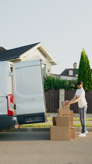 A man dressed in a white t-shirt and jeans is standing beside the open rear doors of a white van, which is parked on a residential street with a house and greenery visible in the background. The man is unpacking cardboard boxes, with two stacked on the ground and one in his hands. The boxes are made of brown corrugated cardboard, some sealed with packing tape. Inside the van, visible packing materials include plastic wrap and protective blankets draped over furniture or other items being loaded or unloaded. The scene depicts the loading or unloading process during a home relocation, with the man preparing items for transport or unpacking at the new location as part of a professional removals service by Man with Van Lambeth. The environment is well-lit, suggesting daytime, and the street has a clean, residential feel with a pavement, fence, and trees nearby.