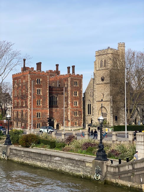 A view of a historic brick castle-like building with battlements and narrow windows, situated on the banks of the River Thames in London. Next to the castle is a tall, square stone church with a clock face and arched windows, reflecting typical medieval architectural features. In the foreground, a paved promenade runs parallel to the river, decorated with black ornate lamp posts, benches, and greenery including neatly trimmed bushes and small trees. Several pedestrians are walking along the promenade, some near the entrance of the church and others along the riverbank. The entire scene is outdoors during daylight, with a partly cloudy sky overhead. This setting captures a typical London riverside area often associated with historical landmarks, ideal for house removals and furniture transport services by companies like Man with Van Lambeth, especially when planning home relocation or packing and moving processes near key sites such as Vauxhall Station and Lambeth Palace.